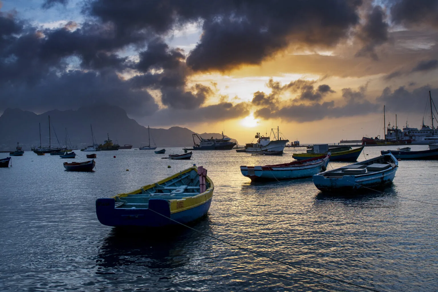 Houten vissersboten dobberen in het water van de haven op São Vicente, verlicht door een sfeervolle zonsondergang met wolken op Kaapverdië.