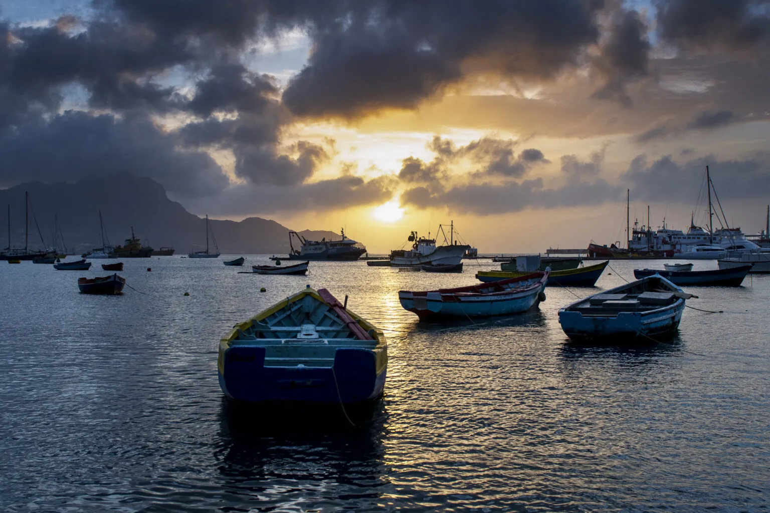 Dobberende vissersbootjes op het water in een Baai van Mindelo terwijl de zon goudgeel ondergaat achter de bergen.