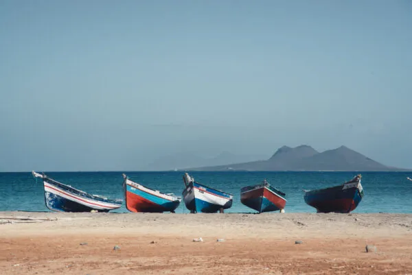 Een rij kleurrijke houten vissersboten op het zandstrand van São Vicente met zicht op de oceaan en bergen op de achtergrond, Kaapverdië.