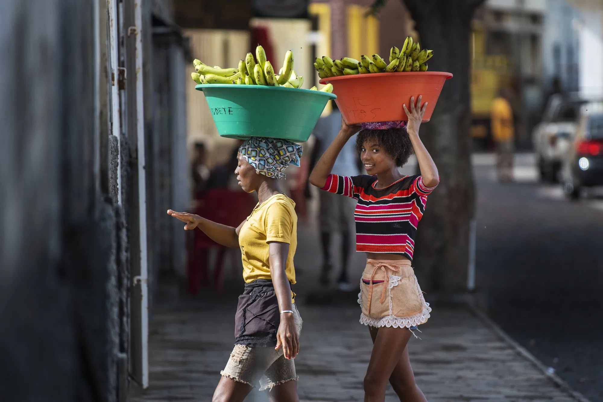 Twee vrolijke jonge lokale vrouwen lopen met grote bakken bananen op hun hoofd op straat op het eiland São Vicente, Kaapverdië