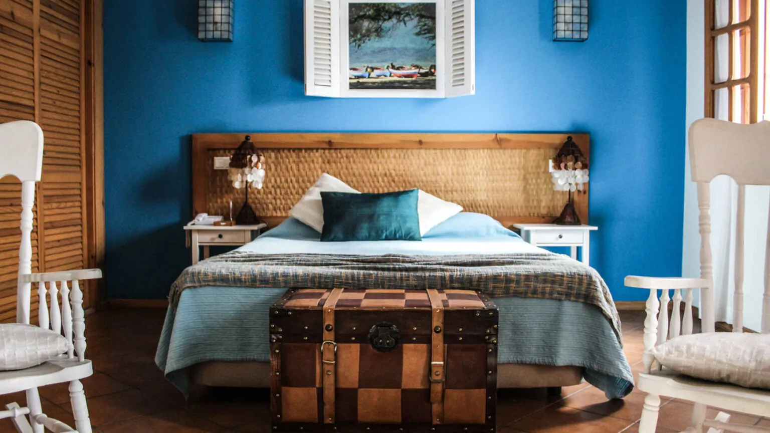 Een sfeervolle hotelkamer met een blauwe muur, een houten tweepersoonsbed, twee witte stoelen en een koffer als salontafel in Kira's Boutique Hotel in Mindelo, Kaapverdië.