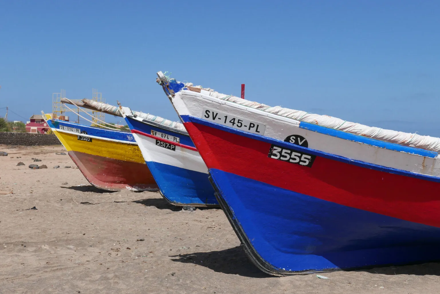 Een rij felgekleurde houten vissersboten ligt op het zandstrand van São Vicente onder een helderblauwe lucht op Kaapverdië.
