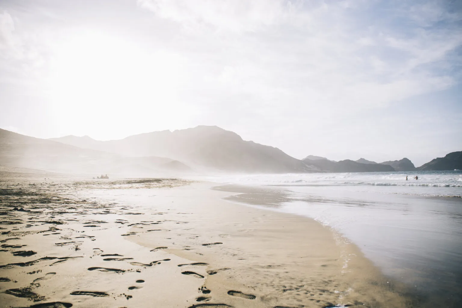 Zandstrand met voetstappen in het natte zand, aangespoelde golven en nevelige bergen op de achtergrond onder een felle zon op het eiland São Vicente in Kaapverdië