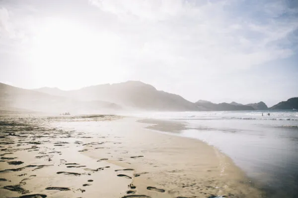 Zandstrand met voetstappen in het natte zand, aangespoelde golven en nevelige bergen op de achtergrond onder een felle zon op het eiland São Vicente in Kaapverdië