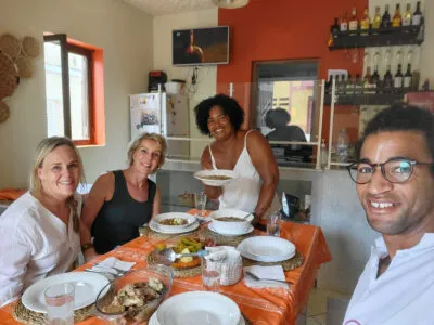 Portret van tante Neusa met twee vrouwelijke reizigers en Voja's lokale gids; samen eten ze de maaltijd die ze hebben bereid tijdens de workshop.