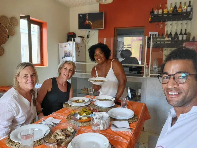 Portret van tante Neusa met twee vrouwelijke reizigers en Voja's lokale gids; samen eten ze de maaltijd die ze hebben bereid tijdens de workshop.