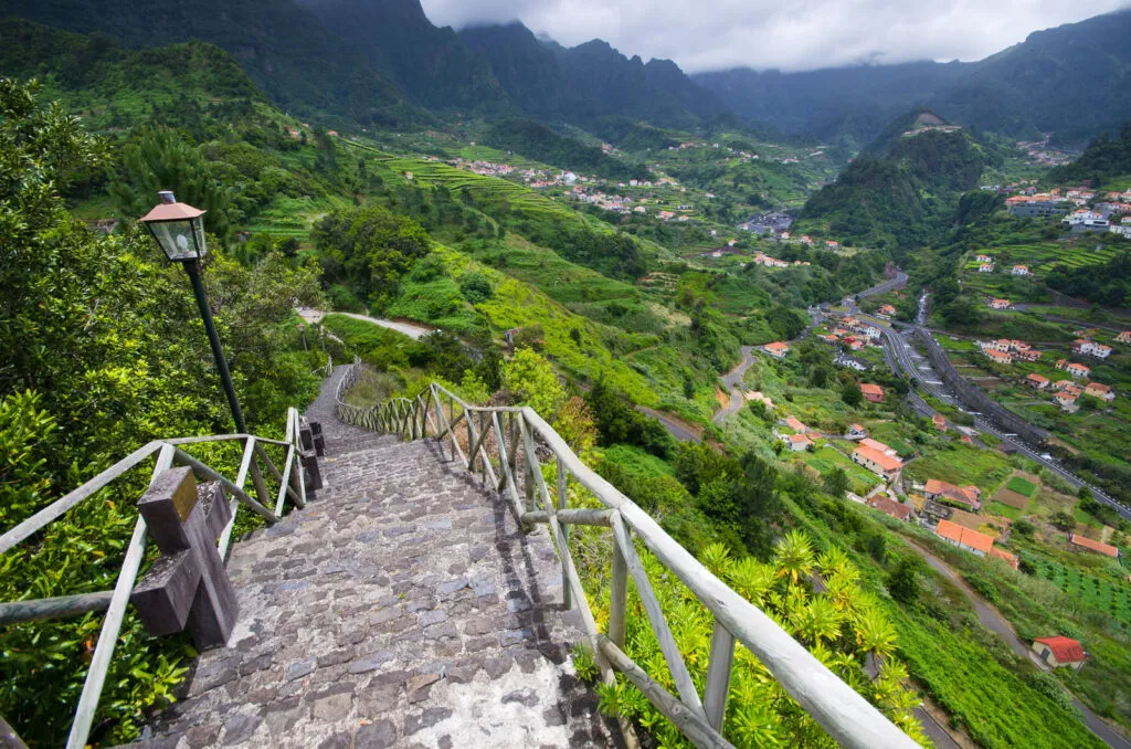 Een smalle stenen trap met leuning leidt naar beneden met uitzicht op een groene vallei, traditionele huizen en steile bergen in São Vicente op Madeira.