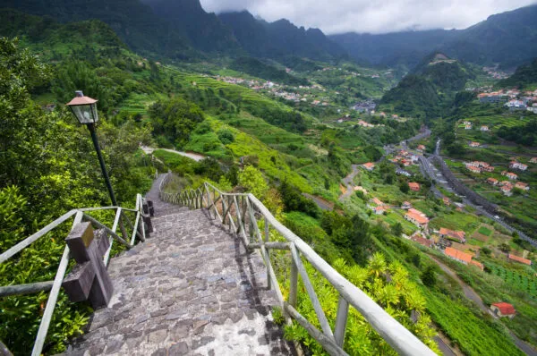 Een smalle stenen trap met leuning leidt naar beneden met uitzicht op een groene vallei, traditionele huizen en steile bergen in São Vicente op Madeira.