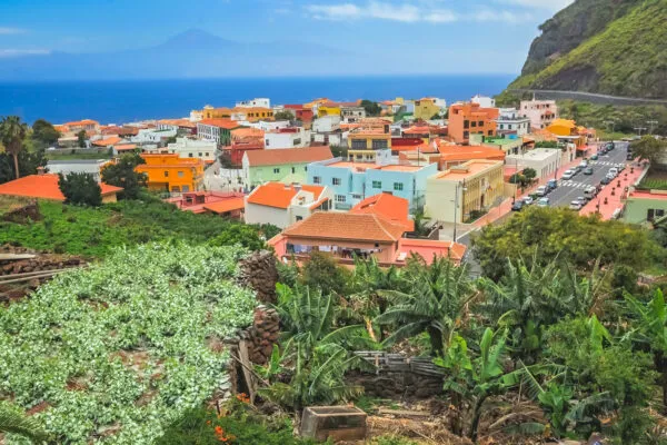 Een dorpsgezicht van Vallehermoso met kleurrijke huizen en oranje daken, omgeven door groene vegetatie en de blauwe oceaan op de achtergrond.