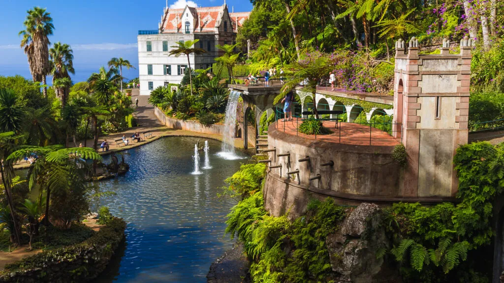 Vijver met spuitende fonteinen en een kleine waterval omgeven door weelderige tropische planten en een historisch landhuis in Monte Palace Garden te Funchal op Madeira