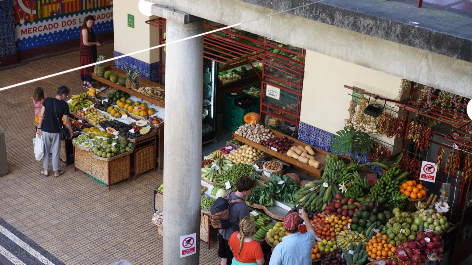 Mensen bekijken een rijke uitstalling van vers tropisch fruit, groenten en bloemen tussen de klassieke stenen pilaren van de markt Mercado dos Lavradores in Funchal op Madeira.