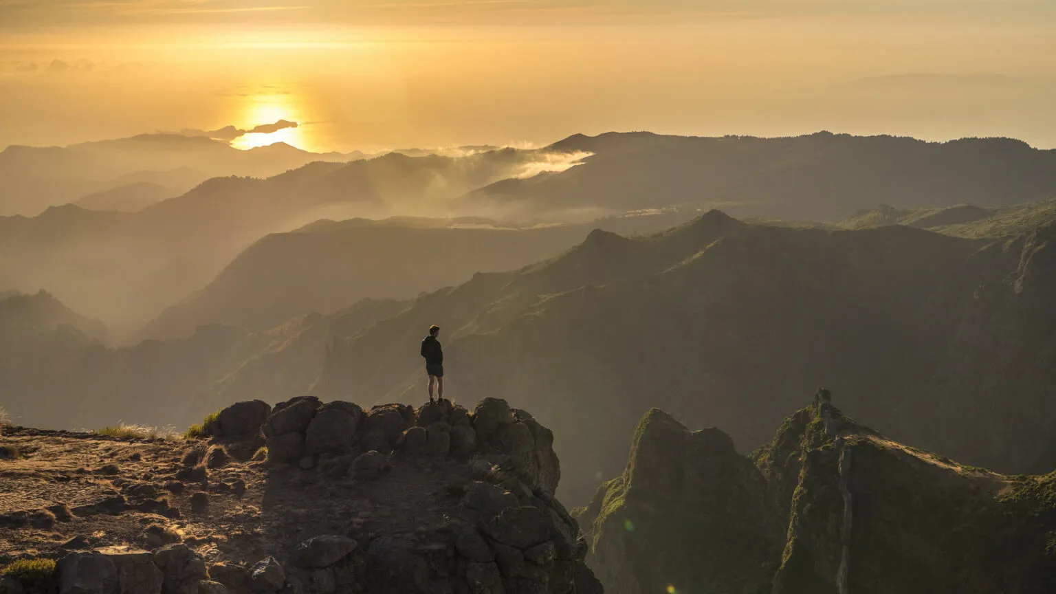 Een reiziger staat op een rotsige uitkijkpost en kijkt uit over de weidse, gelaagde bergketens van Madeira die gehuld zijn in de warme, gouden gloed van de zonsondergang.