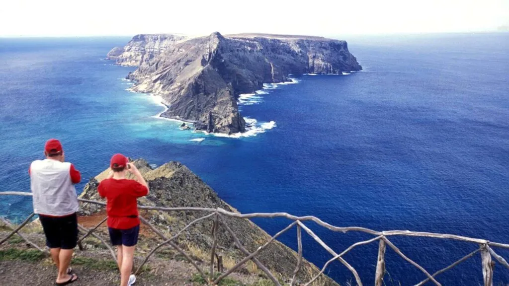 Twee bezoekers kijken vanaf een uitkijkpunt met houten omheining uit over de diepblauwe oceaan en het rotsachtige eilandje Ilhéu de Baixo bij Porto Santo