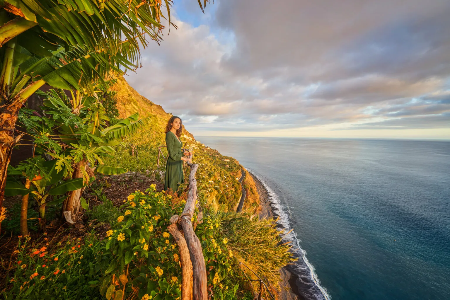 Een vrouw staat bij een houten hekwerk op een groene klif met bananenplanten en kijkt tijdens zonsondergang uit over de Atlantische Oceaan op Madeira.