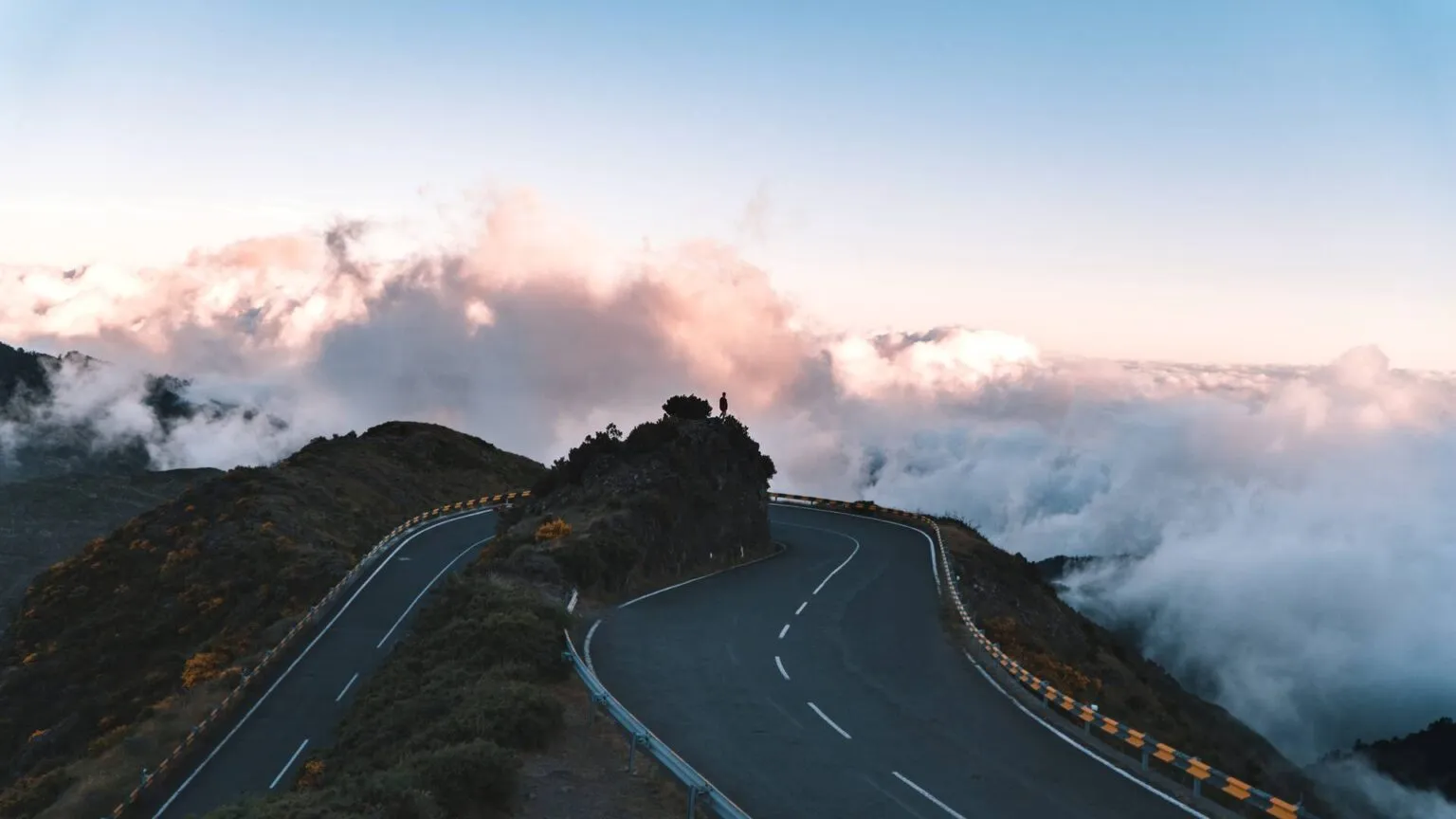 Een kronkelende geasfalteerde autoweg boven de wolken tijdens zonsopgang, met een spectaculair uitzicht over de bergtoppen van Madeira.
