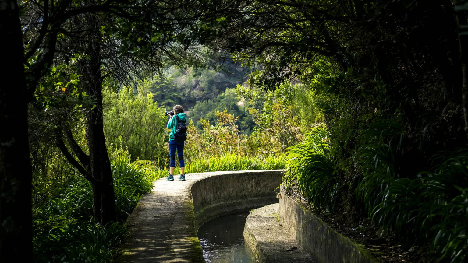 Een wandelaar met rugzak staat stil op het pad langs een smalle levada, omringd door dichte groene bomen en vegetatie op Madeira.