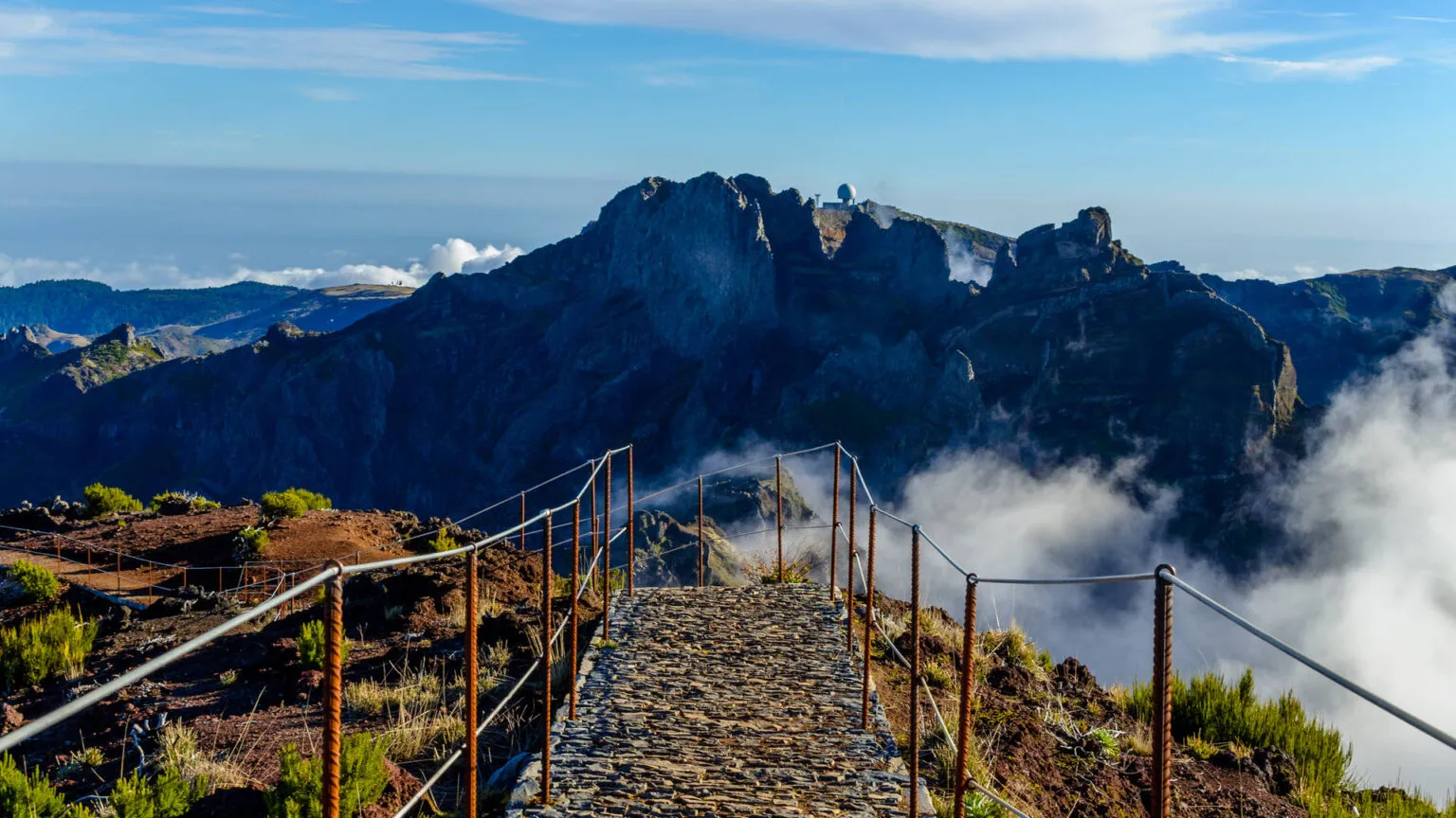 Een geplaveid wandelpad met leuningen leidt langs een bergbepaling richting de imposante rotsen en pieken van Pico Ruivo op Madeira.