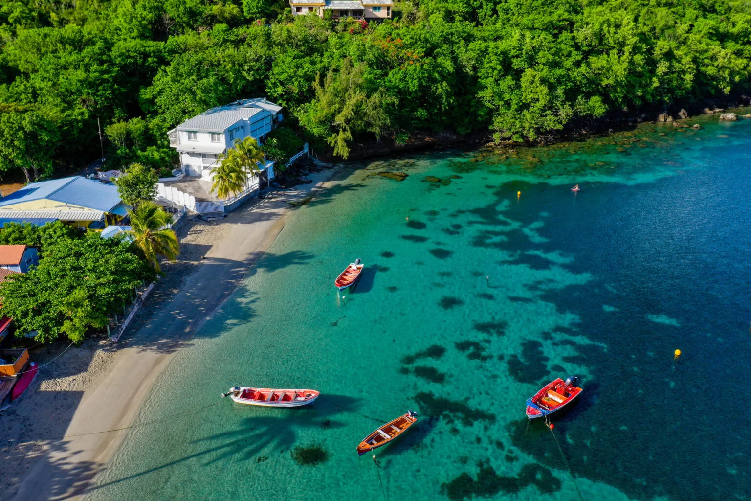 Bovenaanzicht van kleine, kleurrijke houten bootjes die dobberen in een turquoise baai bij een zandstrand en een groen begroeide kust in Grande Anse d'Arlet op Martinique.