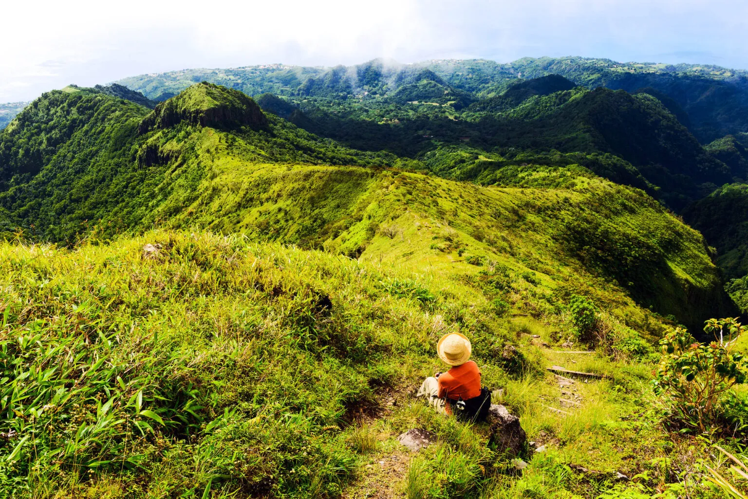 Een hiker in een oranje shirt wandelt over een smal, groen bergpad op de bergkam van de vulkaan Mont Pelée met een panoramisch uitzicht over de heuvels op Martinique.