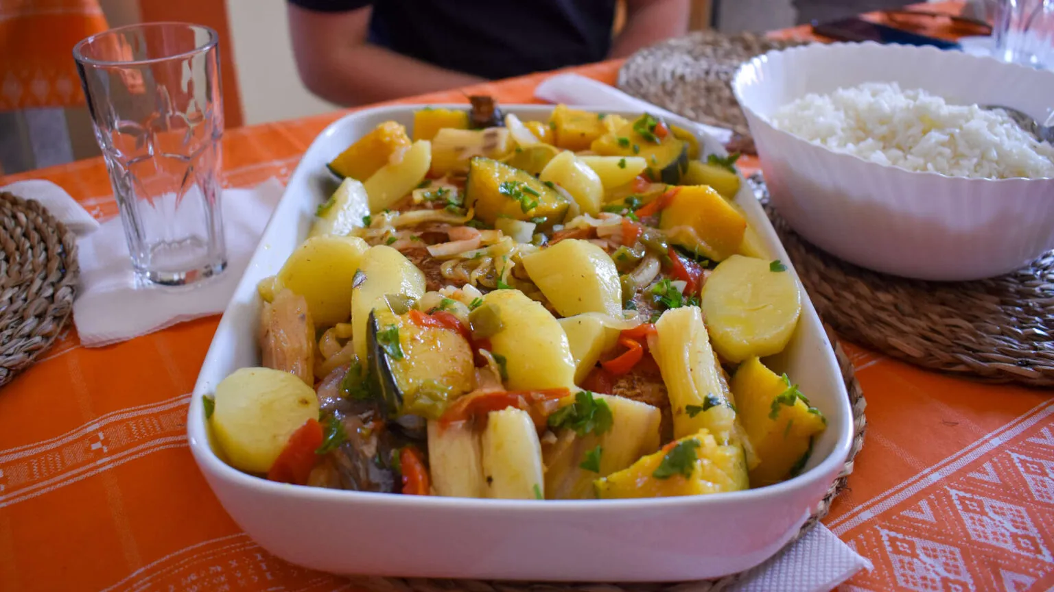 Een witte ovenschaal gevuld met een vers bereide maaltijd van aardappelen, groenten en kruiden op tafel bij Neusa in Kaapverdië.