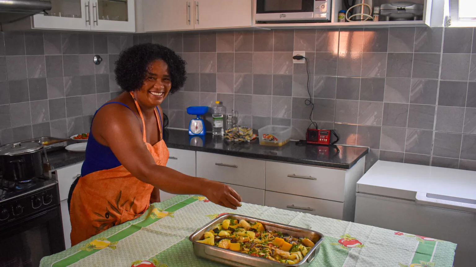 Een glimlachende vrouw genaamd Neusa draagt een oranje schort en kruidt een schaal met groenten op een tafel in een moderne keuken.