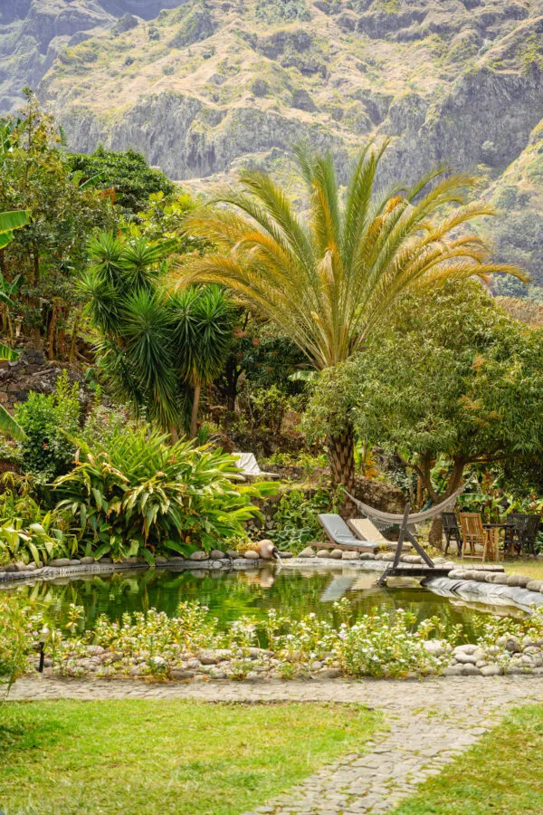 Een weelderige groene tuin met palmbomen, ligstoelen en een zwembad tegen een bergachtige achtergrond bij accommodatie Aldeia Manga op Santo Antão, Kaapverdië.