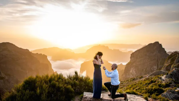 Een man gaat op één knie voor een huwelijksaanzoek aan een vrouw op een bergtop, tegen de achtergrond van een gouden zonsondergang en een wolkendek in het dal.