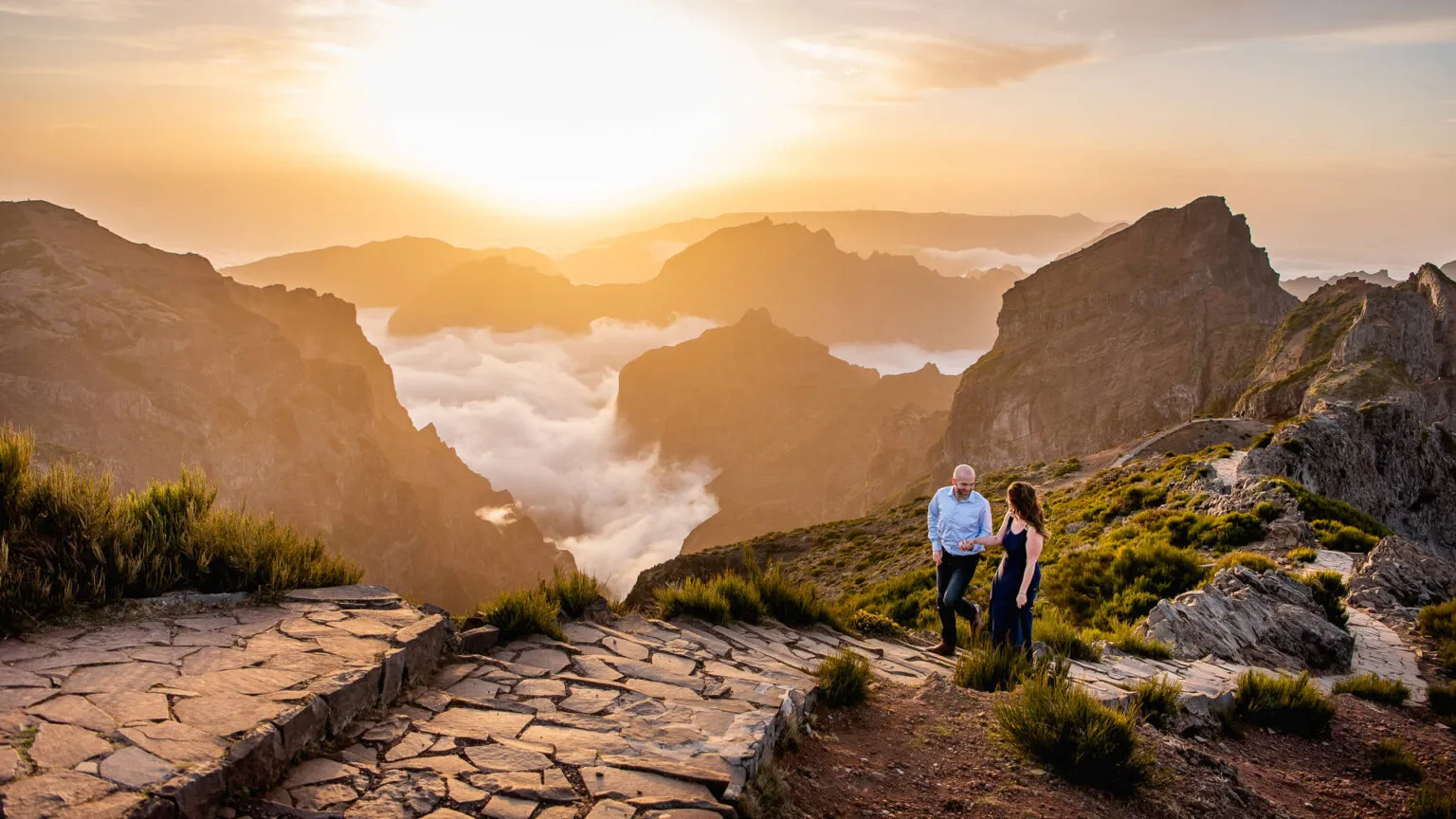 Een man en een vrouw staan samen op een stenen bergpad bij zonsondergang, uitkijkend over de bergtoppen en een deken van wolken in het dal.