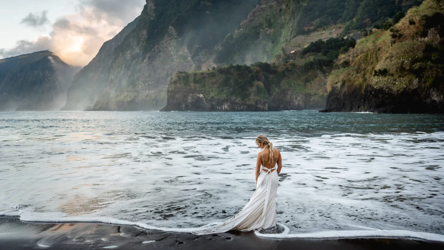 Een bruid in een witte trouwjurk met een lange sleep staat op een donker strand, terwijl het schuimende zeewater rond haar voeten spoelt tegen een decor van steile bergwanden.