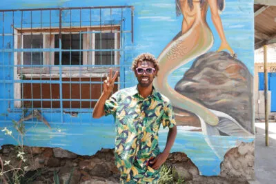 Een vrolijk lachende local steekt twee vingers op voor een muur met kleurrijke streetart in hoofdstad Mindelo van eiland São Vicente