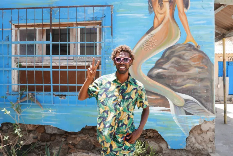 Een vrolijk lachende local steekt twee vingers op voor een muur met kleurrijke streetart in hoofdstad Mindelo van eiland São Vicente