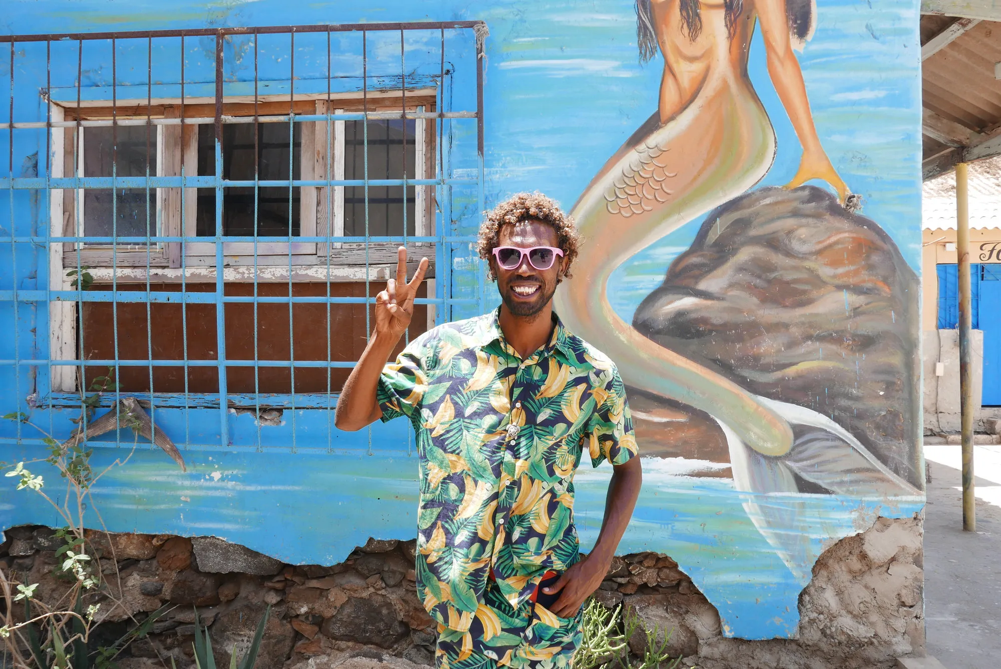 Een vrolijk lachende local steekt twee vingers op voor een muur met kleurrijke streetart in hoofdstad Mindelo van eiland São Vicente