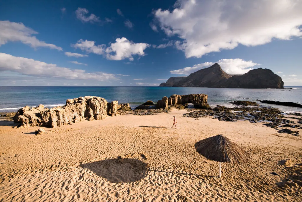 Een goudgeel zandstrand op Porto Santo met rieten parasols op de voorgrond, rotsformaties en uitzicht op de blauwe oceaan onder een lichtbewolkte lucht.