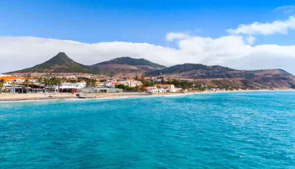 Uitgestrekt goudgeel zandstrand aan de turquoise oceaan met op de achtergrond een kustdorp en vulkanische bergen op Porto Santo.