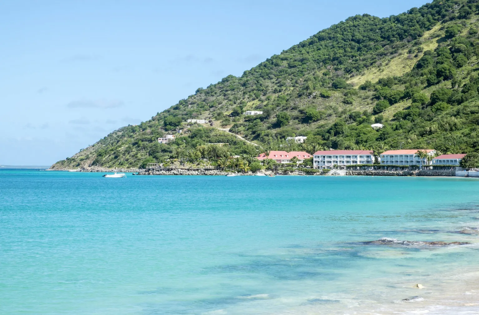 Zicht op het heldere turquoise zeewater van de baai van Grand Case op Saint-Martin, met de gebouwen van de beach club langs de kustlijn en een steile, groenbegroeide berg op de achtergrond.