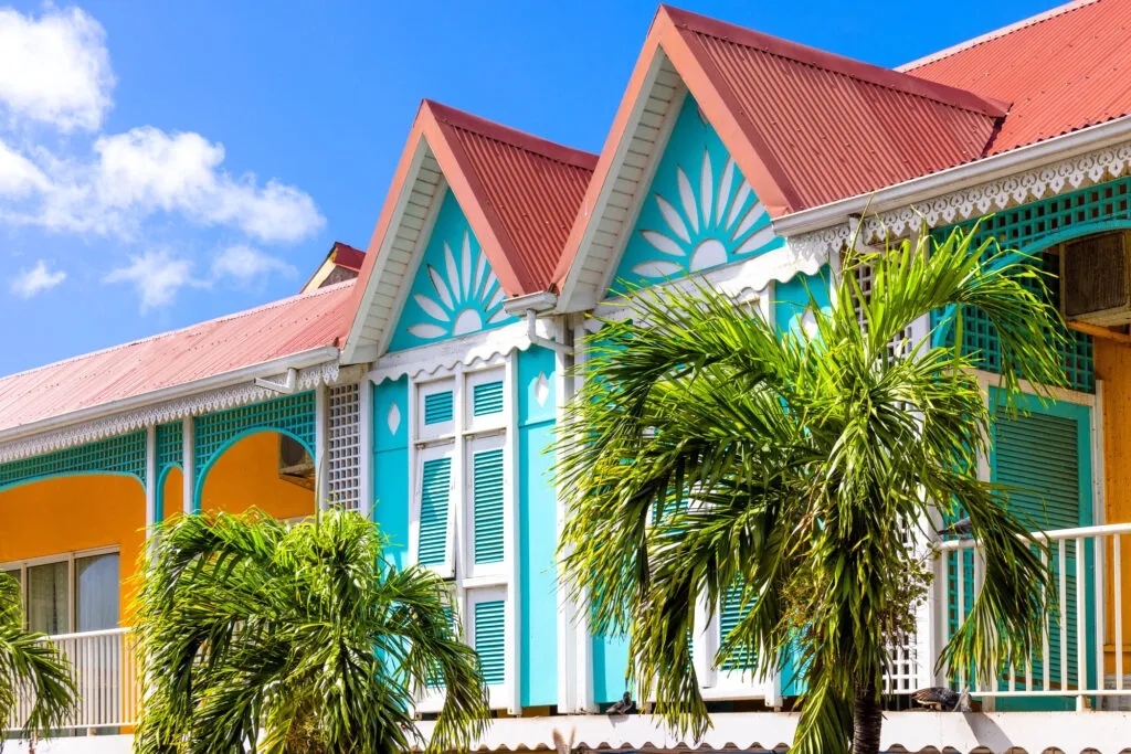 Close-up van een felgekleurd turquoise en geel gebouw met rode puntdaken en houtsnijwerk in Marigot op Saint-Martin, omlijst door groene palmtakken tegen een helderblauwe lucht.