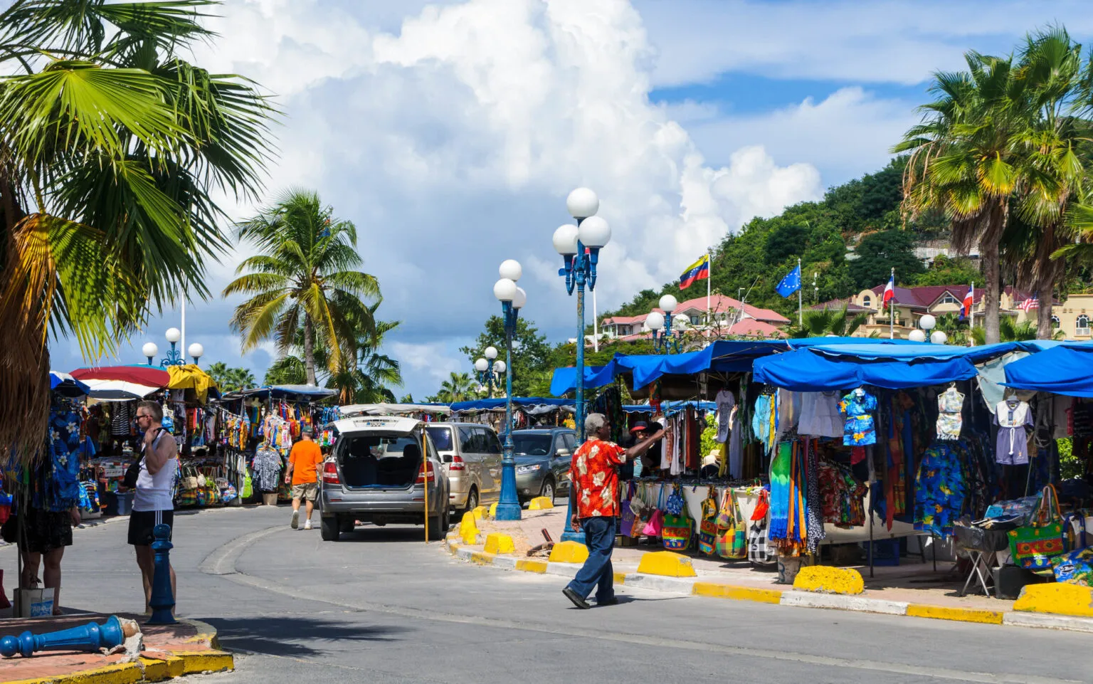 Straatbeeld van een kleurrijke openluchtmarkt op Saint-Martin met diverse marktkramen, wandellende bezoekers en palmbomen tegen een heuvelachtige achtergrond onder een blauwe lucht met wolkpartijen.