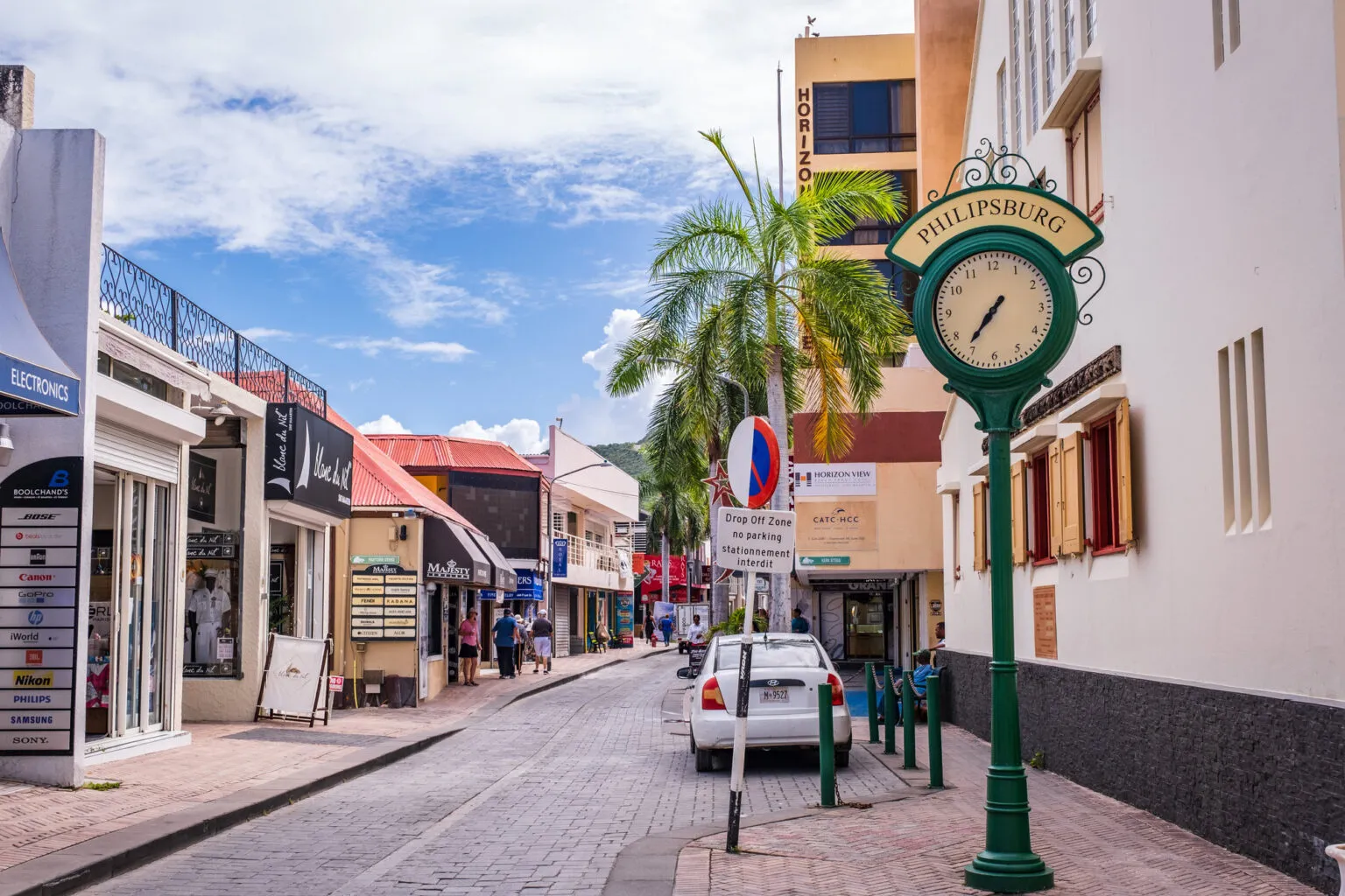 Kleurrijk straatbeeld van een winkelstraat in het centrum van Philipsburg op Sint Maarten, met een klassieke groene klok op een paal op de voorgrond en een palmboom langs de gevels.