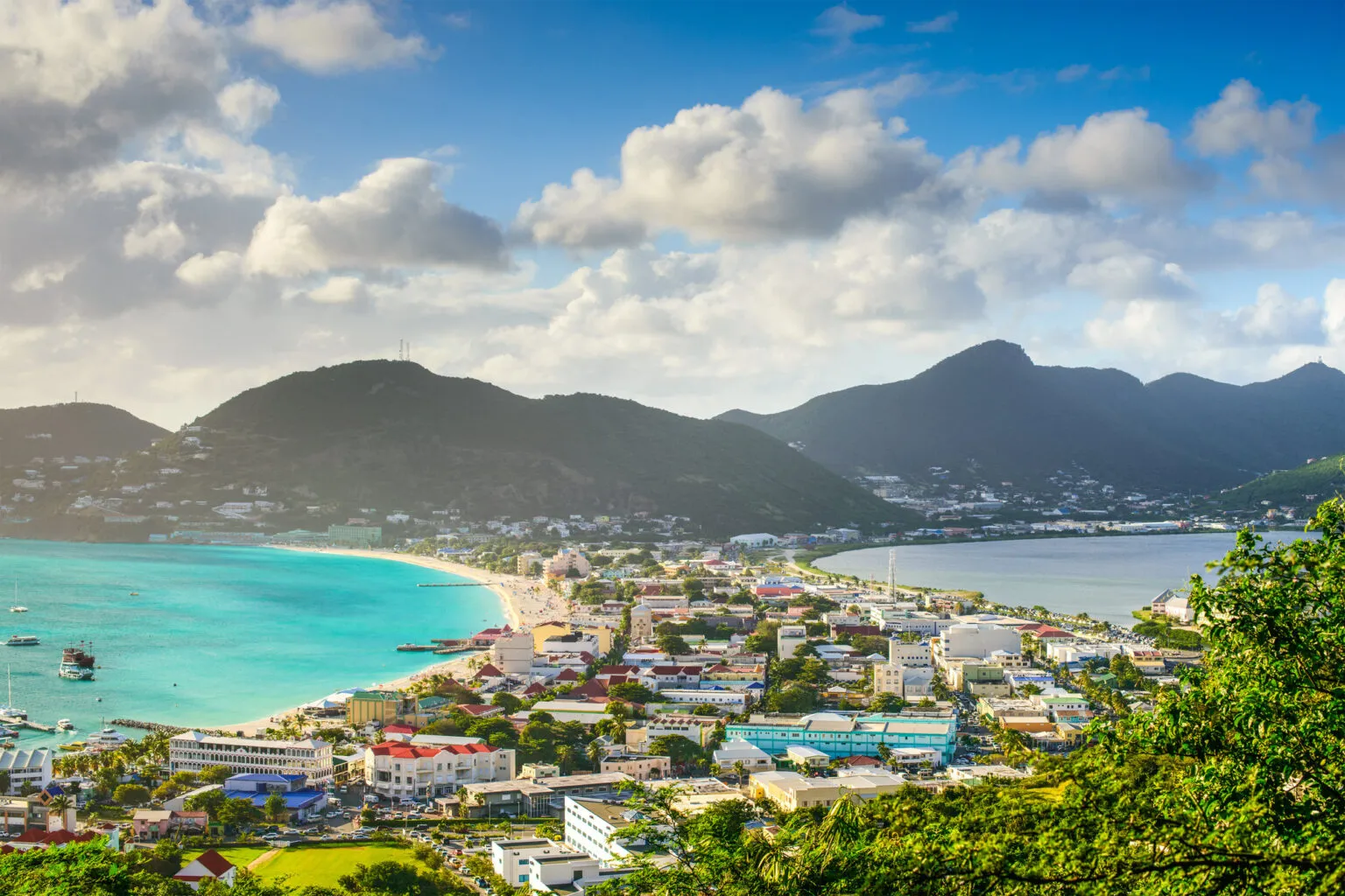 Luchtfoto van Philipsburg op Sint Maarten, met de bebouwde landengte tussen Great Bay met turquoise water links en de zoutpannen rechts, omgeven door groen heuvellandschap onder een lichtbewolkte blauwe lucht.