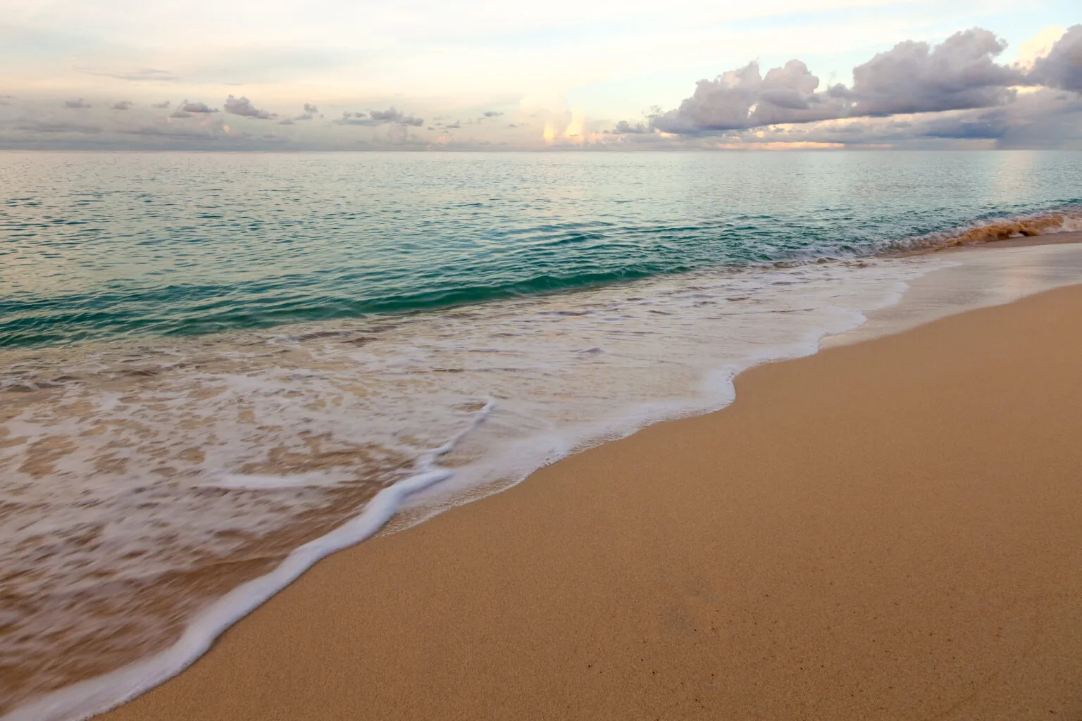 Lichte golfslag die op een goudgeel zandstrand spoelt met op de achtergrond een rustige zee en bewolkte lucht tijdens zonsopgang op Sint-Maarten.