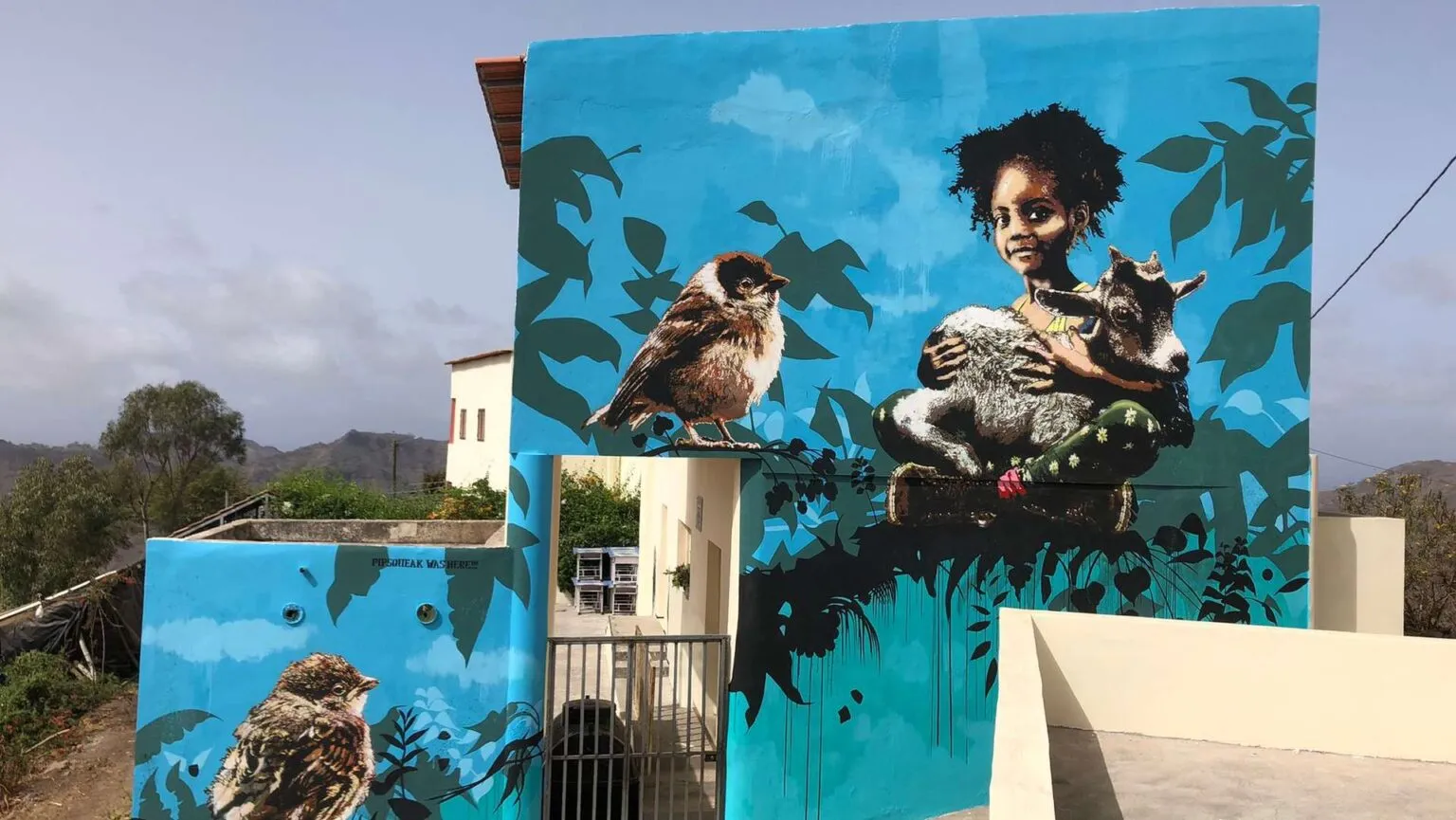 Een kleurrijke blauwe muurschildering op een gebouw toont een kind dat een geitje vasthoudt naast een grote vogel in Fundura, Santiago.