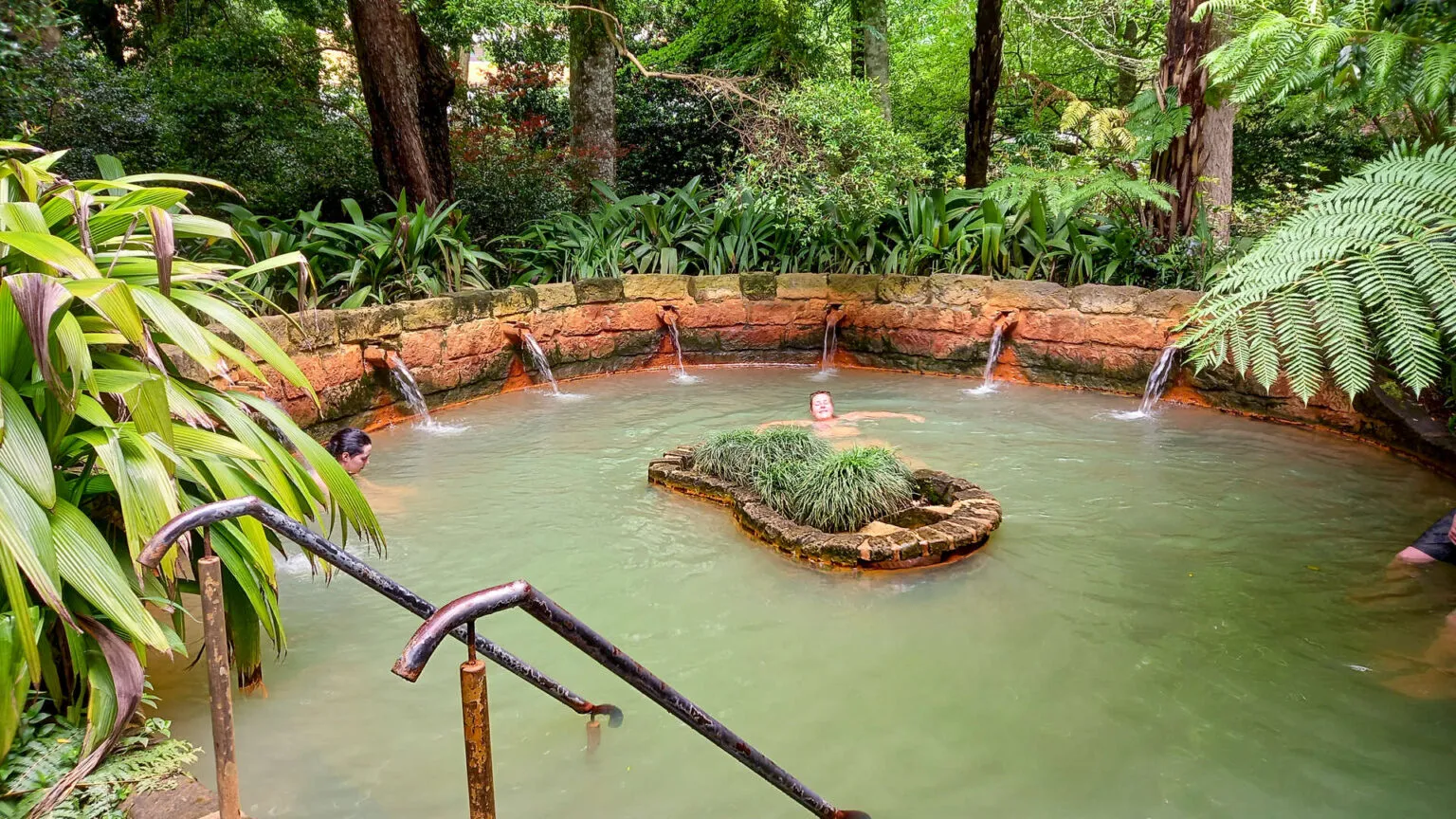 Het groenachtige water van het grote geothermische bad in Caldeira Velha op São Miguel met een klein eilandje in het midden en een trap op de voorgrond.