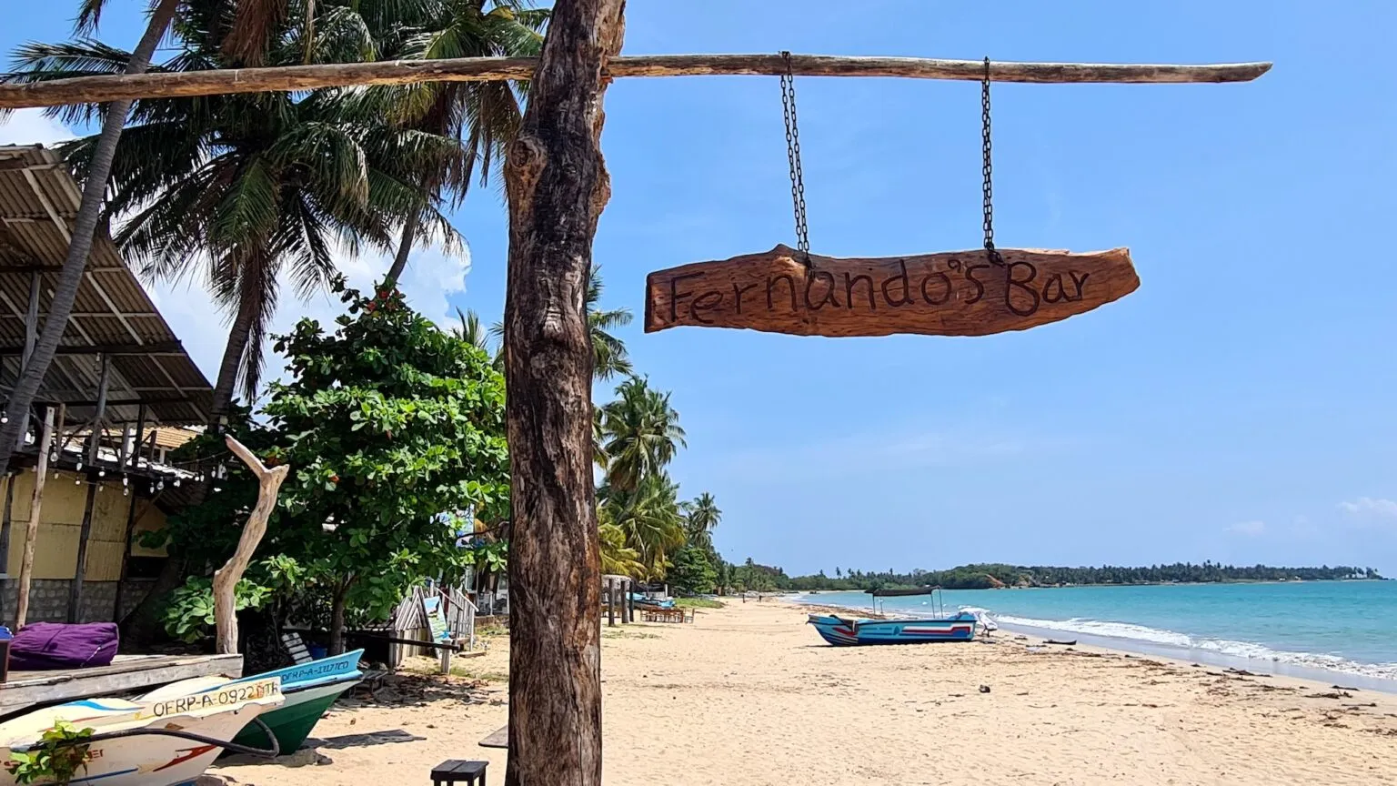 Een houten naambord van Fernando's Bar hangt aan een paal op een zandstrand in Uppuveli, met vissersboten en een helderblauwe zee op de achtergrond.