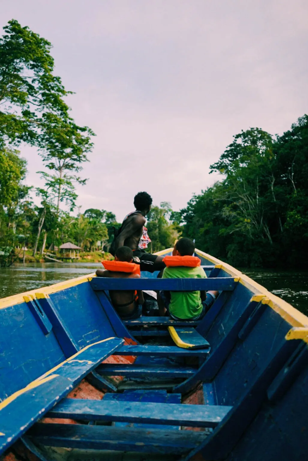 Perspectief vanaf de achterkant van een houten korjaal-boot met een man en kinderen over een rivier omgeven door dicht tropisch regenwoud in Suriname.