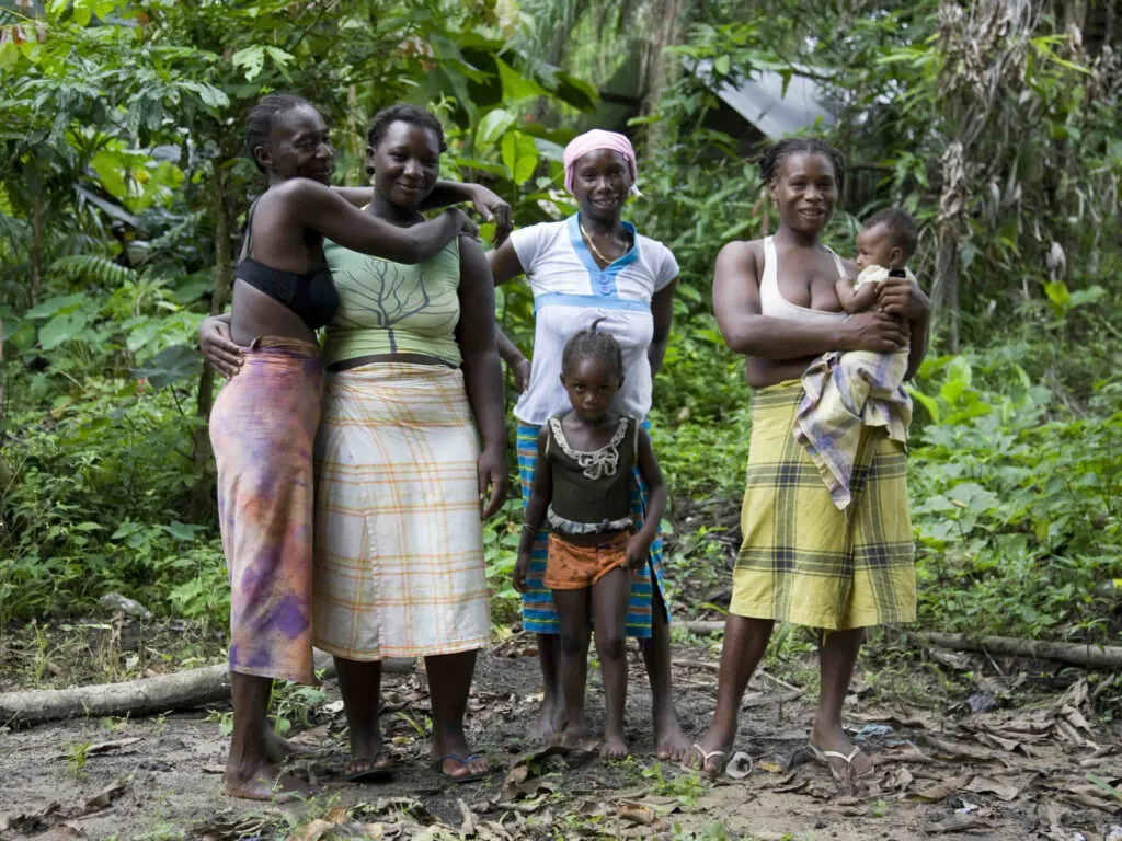 Een groep Surinaamse Marronvrouwen en kinderen gepositioneerd voor een groen woudlandschap, gekleed in kleurrijke pangi-doeken.