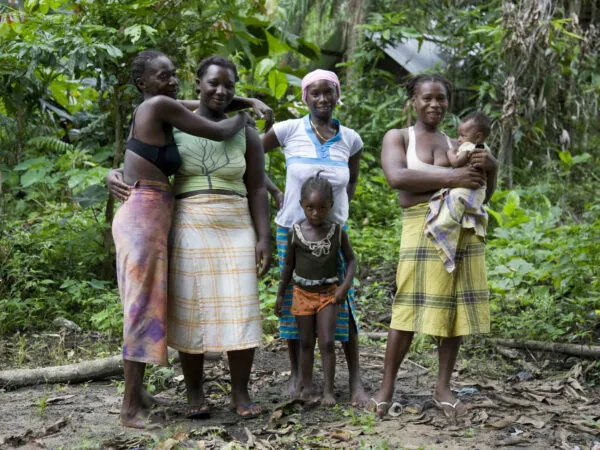 Een groep Surinaamse Marronvrouwen en kinderen gepositioneerd voor een groen woudlandschap, gekleed in kleurrijke pangi-doeken.