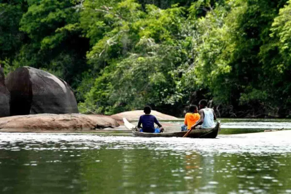 Aanzicht vanaf de achterzijde van een houten korjaal met inzittenden die over een rustige rivier varen, langs rotsen en dichtbegroeid tropisch oerwoud in Suriname.