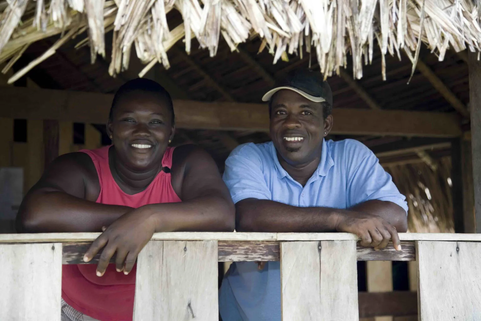 Portret van een glimlachende Surinaamse man en vrouw die leunen op de houten balustrade van een traditionele woning met een rieten dak.
