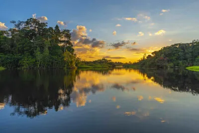 De ondergaande zon verlicht de wolken boven een brede rivier in Suriname, met spiegelingen in het kalme water en dicht begroeide tropische jungle aan de oever.