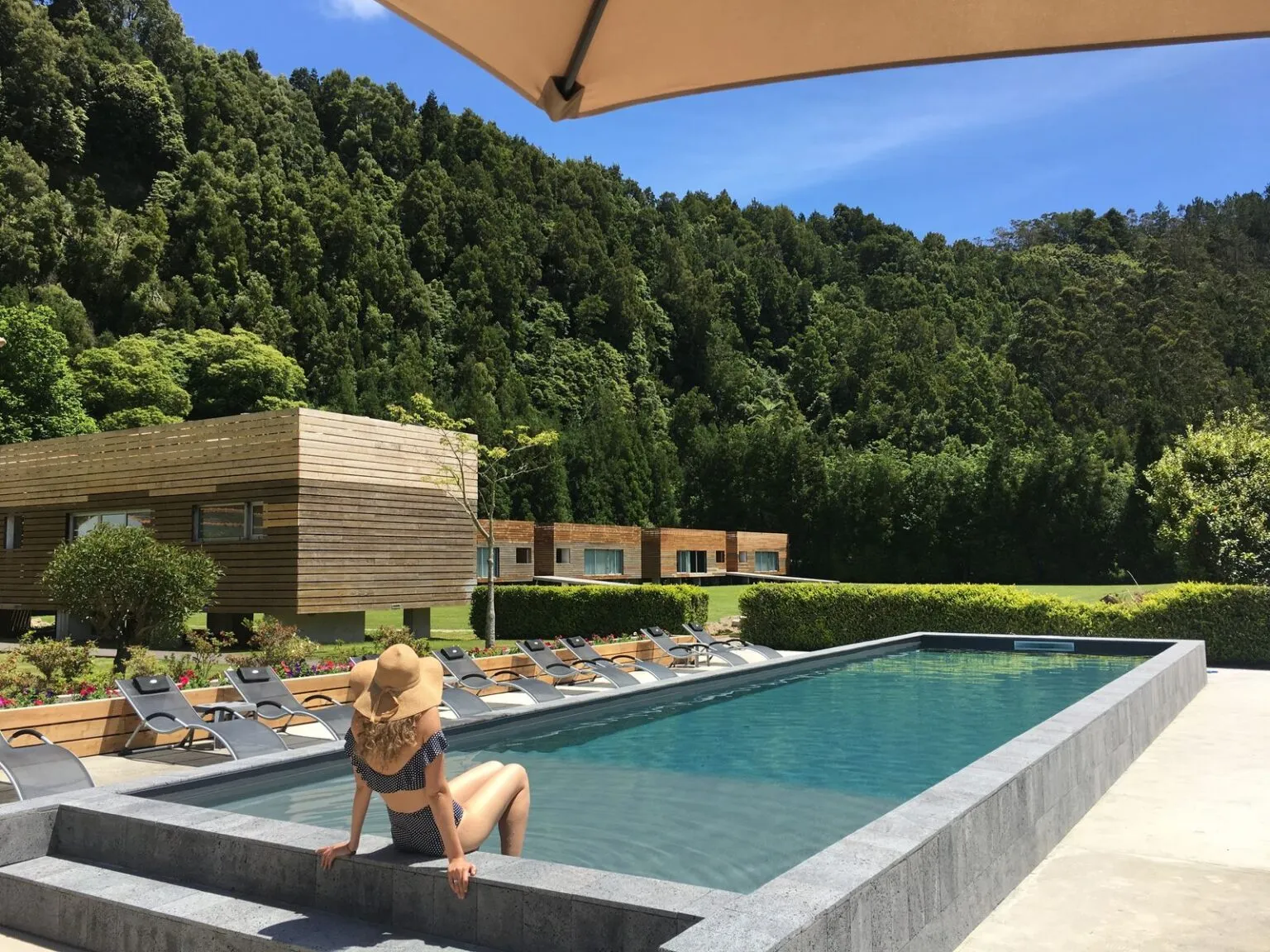 Een vrouw zit aan de rand van een strak zwembad, omgeven door een strakke tuin en dicht begroeide groene heuvels bij Furnas Lake Forest Living.